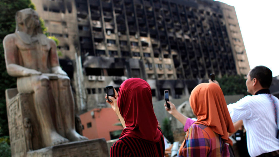 Туристы фотографируют статую перед&nbsp;Каирским музеем на&nbsp;фоне сгоревшего здания правящей партии Египта на&nbsp;площади Тахрир, 2011 год