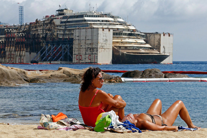 Девушки на пляже острова Джильо на фоне поднятого лайнера Costa Concordia, готовящегося к транспортировке на утилизацию. 21 июля 2014 года
