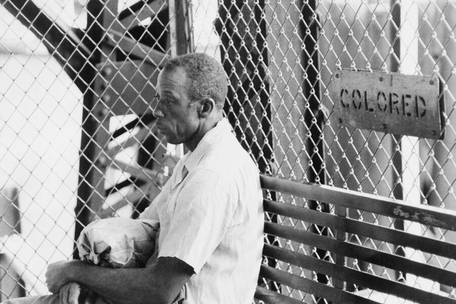 Чернокожий мужчина на фоне сетчатого ограждения, на котором висит табличка «цветной». США, около 1955 года